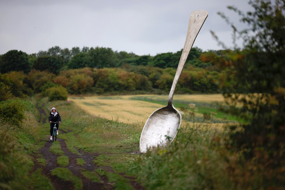 A person cycles past a large sculpture of a spoon on the edge of a field