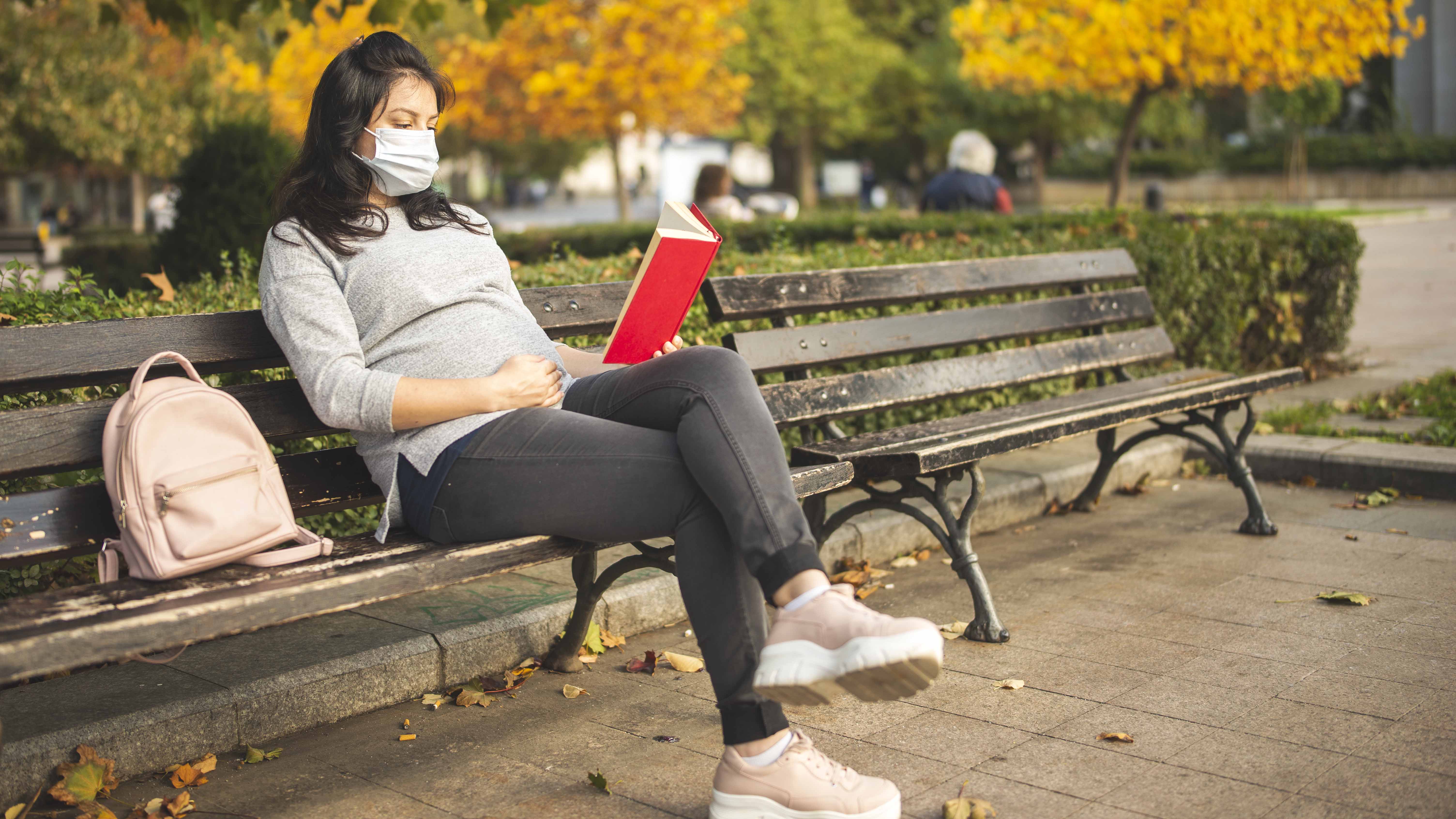 Young pregnant woman with protective face mask reading book sitting on a bench in city park in autumn.