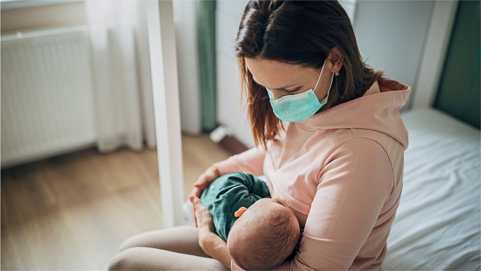 Two people, mother with protective mask breastfeeding her baby son at home