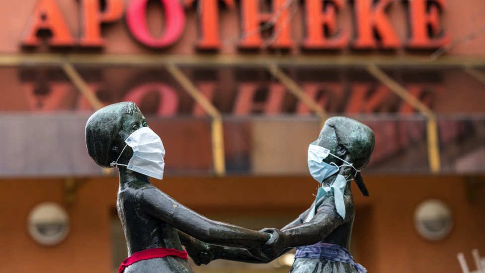 A street sceen in Germany - a statue of two children playing in front of a pharmacy, the statues have been dressed with face masks