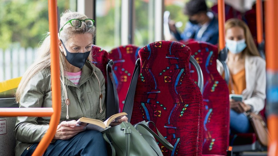 Woman sitting on a bus wearing a protective mask during the Covid 19 pandemic. She is reading a book. There are other passengers behind her who are not in focus.