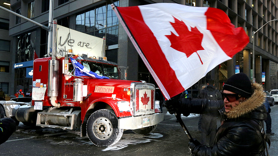 Korona virus, protesti i Kanada: „Psihološki rat" - u Otavi vanredno stanje, sudija naredio kamiondžijama da prestanu da trube 3 A person waves a Canadian flag in front of a truck